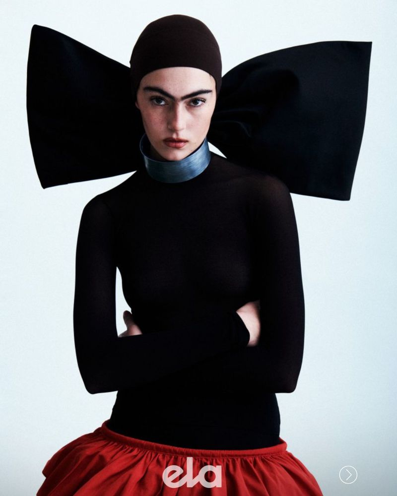 Model wearing a fitted black top, red skirt, dark cap, and large black bow accessory against a plain white background.