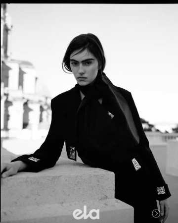 Model in black jacket with patterned embellishments leaning on a stone structure outdoors