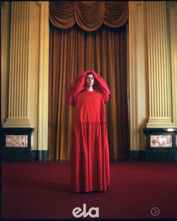 Model wearing a floor-length red hooded gown standing in a grand hall with heavy gold curtains and columns.