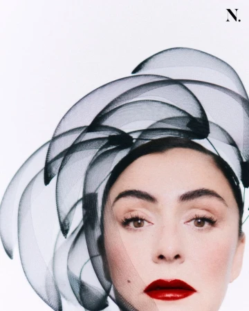 Close-up portrait of a woman wearing an avant-garde black mesh sculptural headpiece and bold red lipstick against a white background.