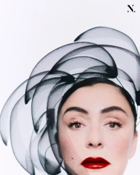Close-up portrait of a woman wearing an avant-garde black mesh sculptural headpiece and bold red lipstick against a white background.