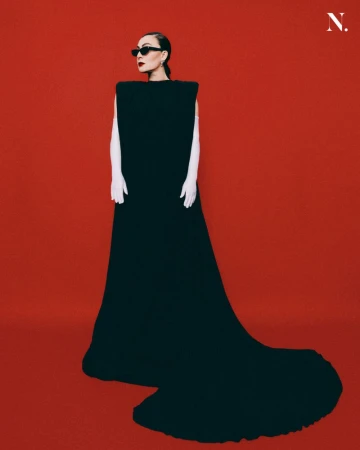 Woman in a structured black gown with white gloves and dark sunglasses standing against a solid red background for Noir Magazine