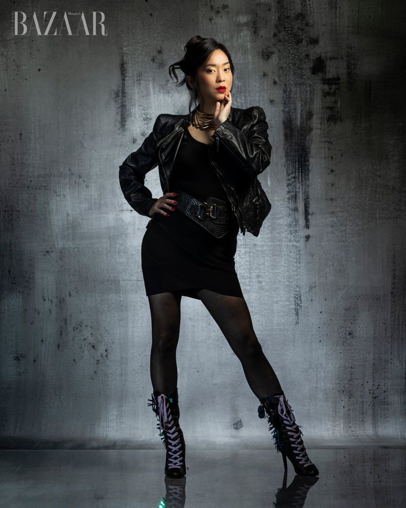 Woman in black leather jacket and dress standing against a textured grey background with Harper's Bazaar logo at top left.