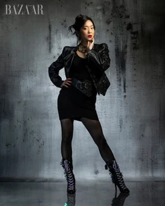 Woman in black leather jacket and dress standing against a textured grey background with Harper's Bazaar logo at top left.