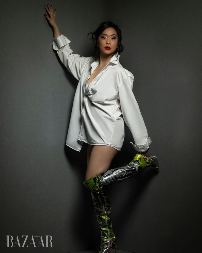 Woman in an oversized white shirt and tall metallic boots posing against a dark grey corner wall.