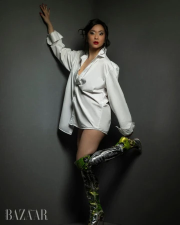 Woman in an oversized white shirt and tall metallic boots posing against a dark grey corner wall.