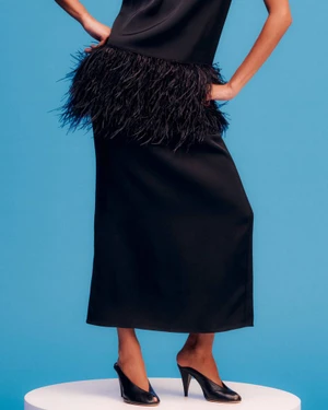 Model wearing a black feather-trimmed dress and black mules standing on a white pedestal against a blue background