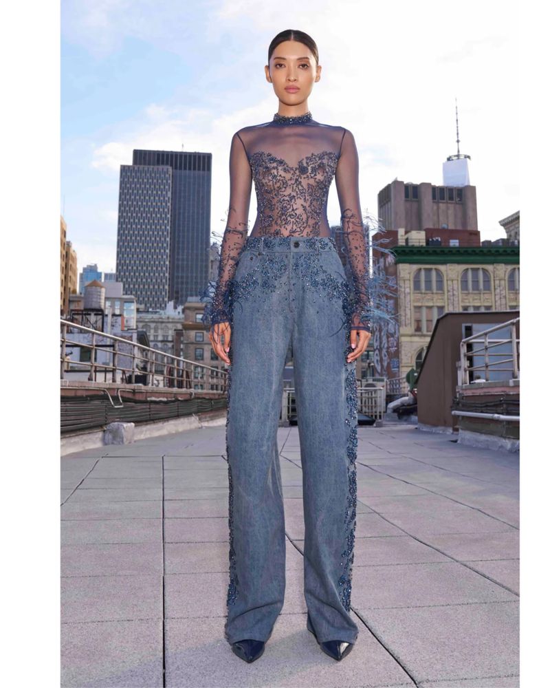 Model wearing a sheer embellished top and wide-leg denim trousers standing on a rooftop overlooking a city skyline.