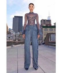Model wearing a sheer embellished top and wide-leg denim trousers standing on a rooftop overlooking a city skyline.