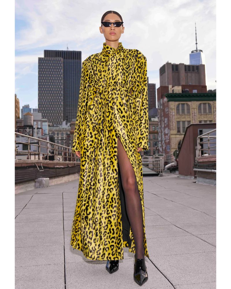 Model wearing a yellow and black leopard print long coat and sunglasses on a rooftop with city skyline backdrop.