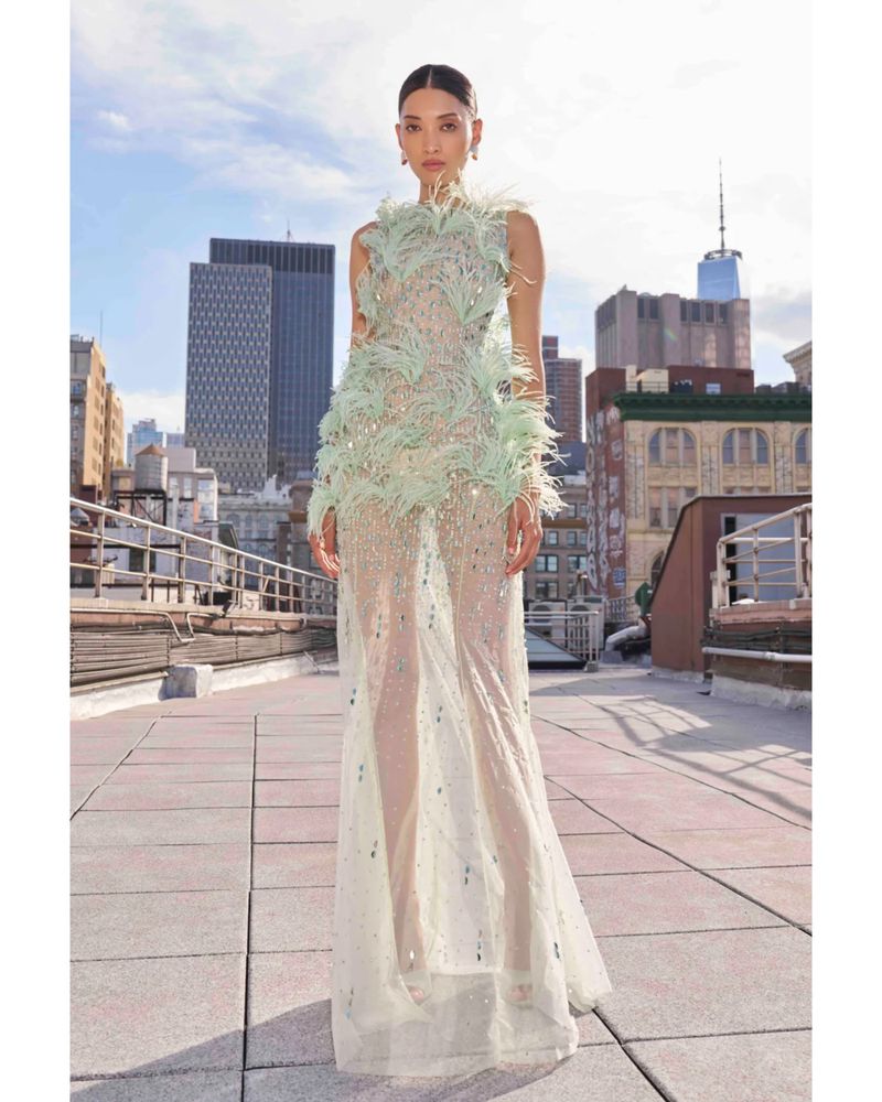 Model wearing a sheer mint green feather-embellished gown standing on an urban rooftop with city skyscrapers in the background.
