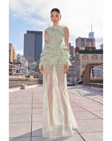 Model wearing a sheer mint green feather-embellished gown standing on an urban rooftop with city skyscrapers in the background.