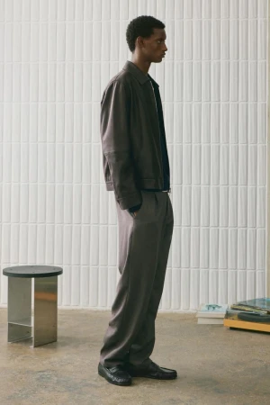 Man wearing a brown leather jacket and grey trousers standing in a minimalist room with a tiled wall