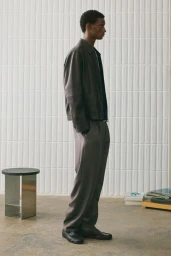 Man wearing a brown leather jacket and grey trousers standing in a minimalist room with a tiled wall
