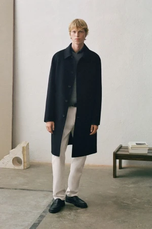 Male model in a black wool coat, grey sweater, and white trousers standing in a minimalist interior space.