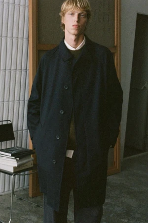 A male model standing in an indoor studio setting wearing a dark coat over layered sweaters.