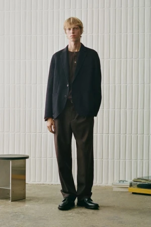 Full-length portrait of a male model wearing a dark blazer, brown trousers, and black shoes against a tiled wall.