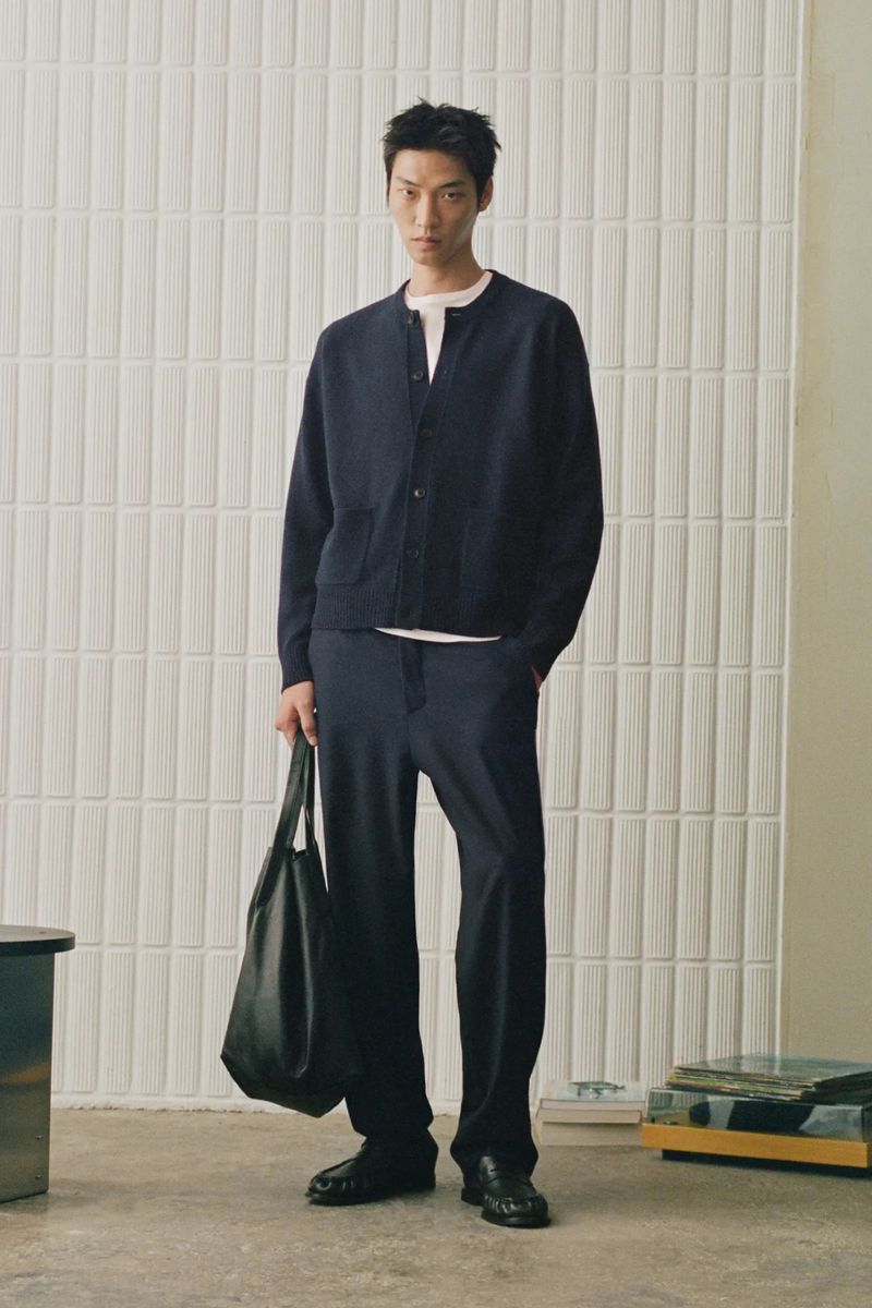 Man wearing a dark cardigan and trousers holding a black tote bag against a textured wall