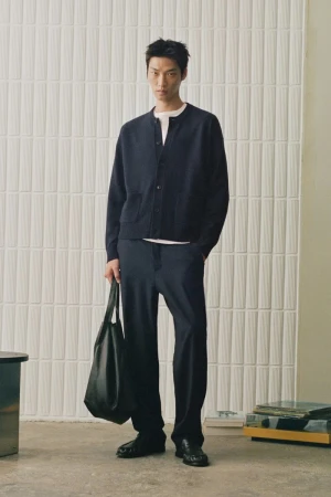 Man wearing a dark cardigan and trousers holding a black tote bag against a textured wall