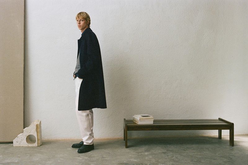 Model standing in a long dark coat and white trousers next to a wooden bench against a textured interior wall.