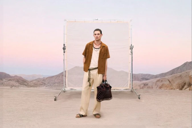 Model standing in front of a photography diffusion screen in a desert landscape wearing cream trousers and a brown shirt