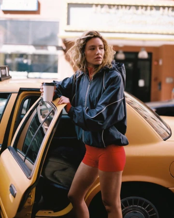 Woman in dark zip-up jacket and red shorts holding a takeaway cup by a yellow taxi