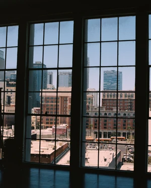 Large multi-pane industrial windows framing an urban view of downtown skyscrapers and historic brick buildings under a clear sky.