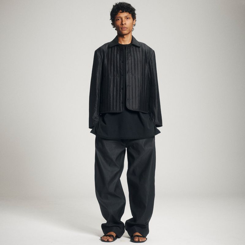 A model poses in a black quilted jacket, dark top, baggy pants, and sandals against a neutral background in a full-length fashion shot.