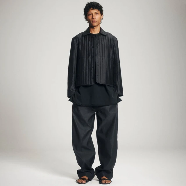 A model poses in a black quilted jacket, dark top, baggy pants, and sandals against a neutral background in a full-length fashion shot.