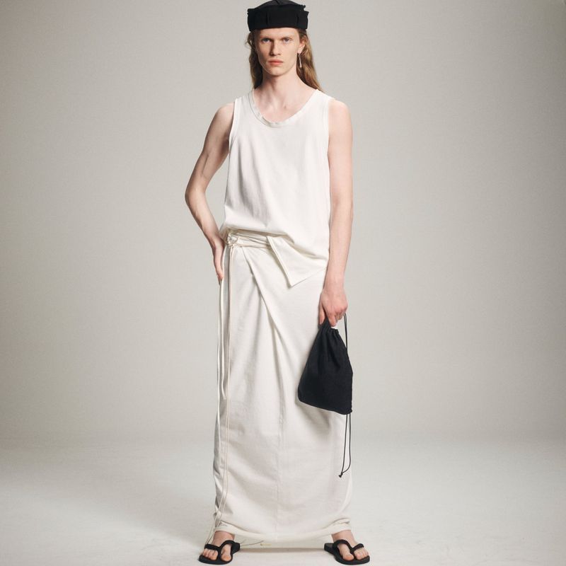 A slender adult male poses in a white tank top and matching long wraparound skirt, accessorized with black sandals and a small bag.