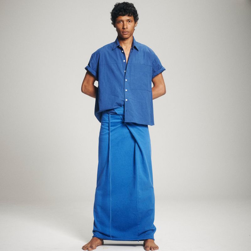 Full shot of a person in a royal blue button-down shirt and matching sarong-style wrap standing against a neutral studio backdrop.