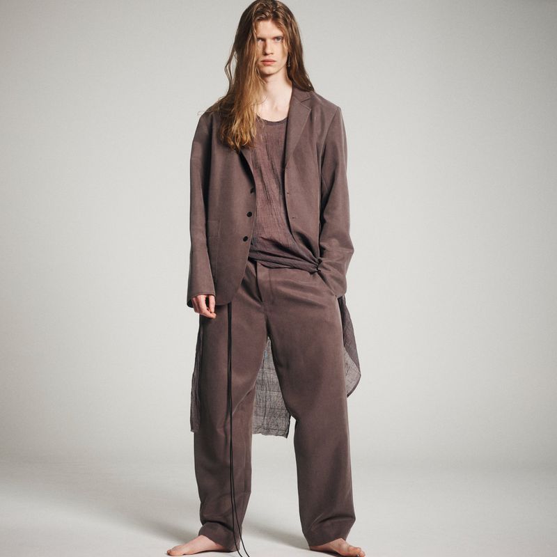 Fashion portrait of a male model in a long brown blazer and pants against a neutral backdrop in a studio setting with soft lighting.