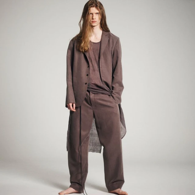 Fashion portrait of a male model in a long brown blazer and pants against a neutral backdrop in a studio setting with soft lighting.