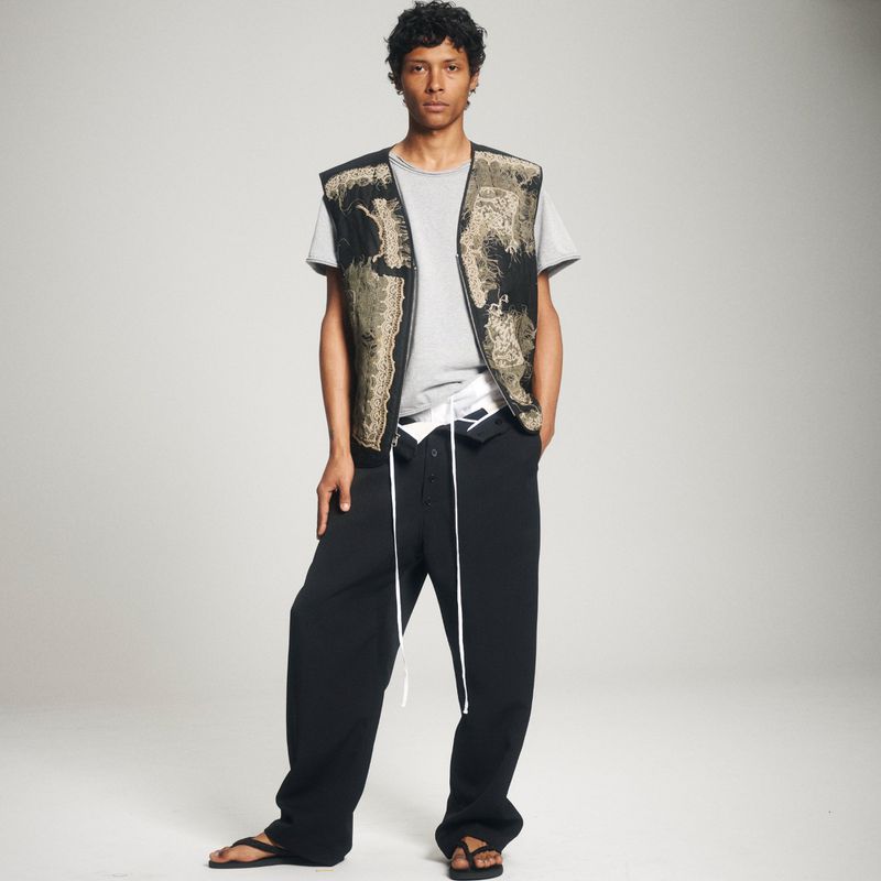 Male fashion model poses in a gray tee, patterned vest, and black pants against a neutral background in a studio fashion shot.