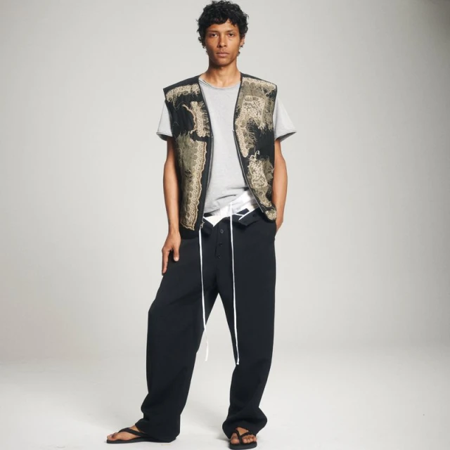 Male fashion model poses in a gray tee, patterned vest, and black pants against a neutral background in a studio fashion shot.