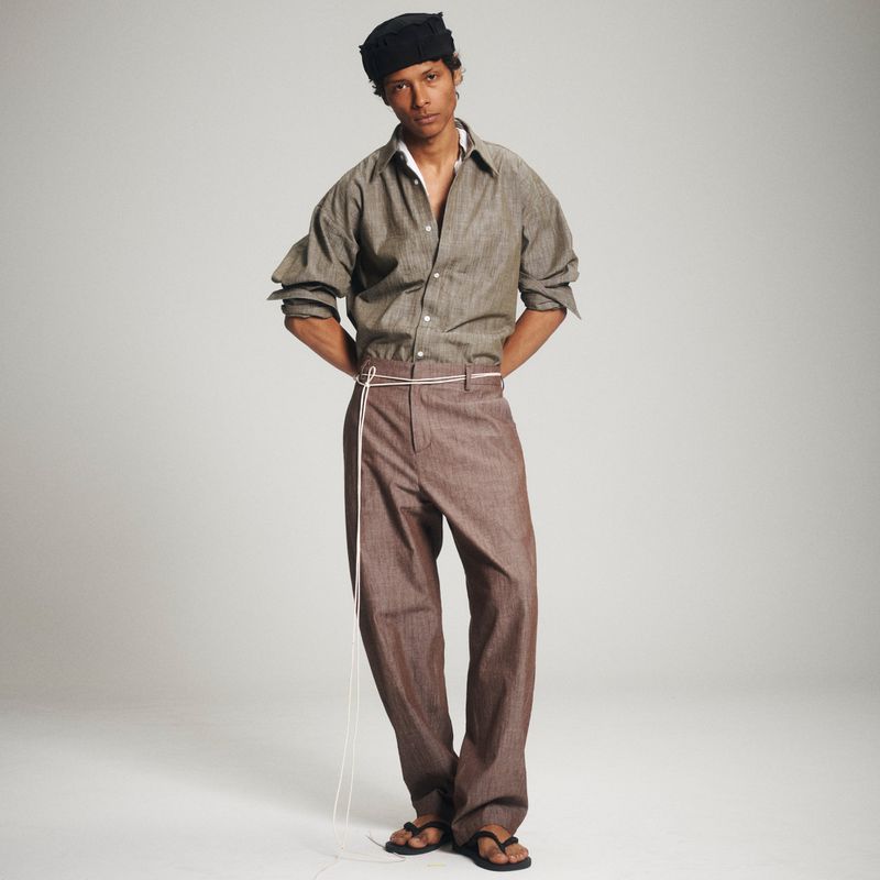 Full-length shot of an adult male fashion model with dark hair wearing a brown shirt and pants, standing against a light grey backdrop.