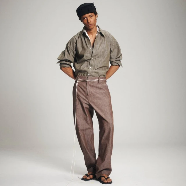 Full-length shot of an adult male fashion model with dark hair wearing a brown shirt and pants, standing against a light grey backdrop.