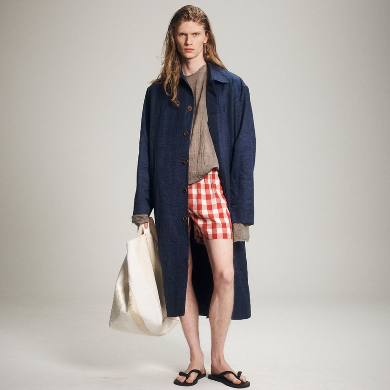 A male model wearing a blue jacket, red and white checkered shorts, and sandals stands against a plain background while holding a tote bag.