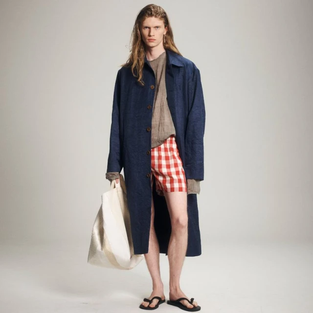 A male model wearing a blue jacket, red and white checkered shorts, and sandals stands against a plain background while holding a tote bag.