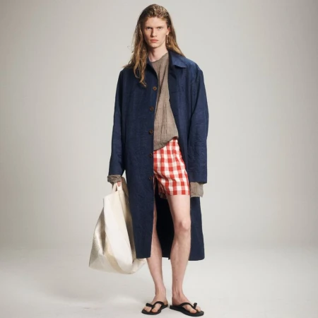 A male model wearing a blue jacket, red and white checkered shorts, and sandals stands against a plain background while holding a tote bag.
