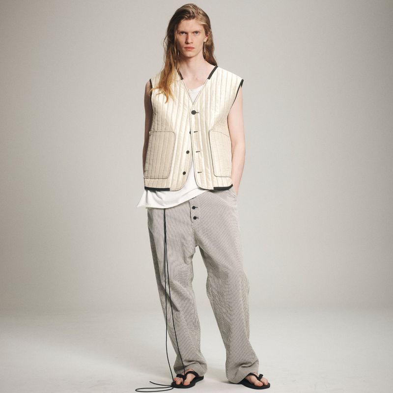 Fashion shot of a slender, fair-skinned adult male model with long blond hair wearing a cream quilted vest and striped trousers in a studio.