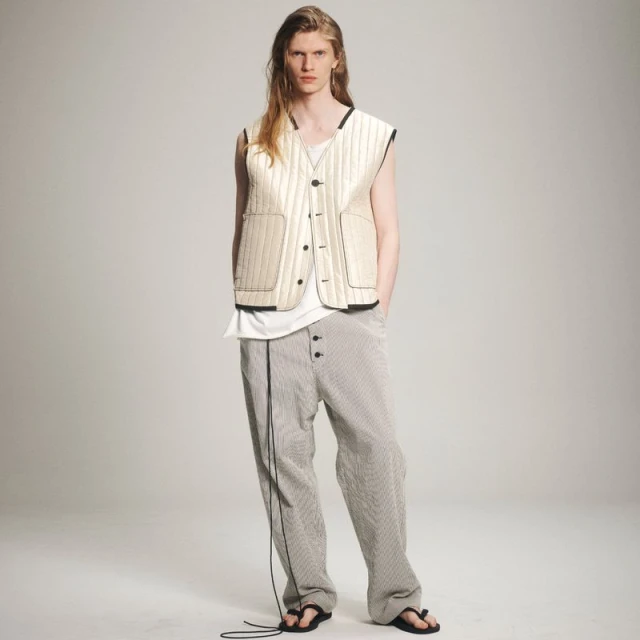 Fashion shot of a slender, fair-skinned adult male model with long blond hair wearing a cream quilted vest and striped trousers in a studio.