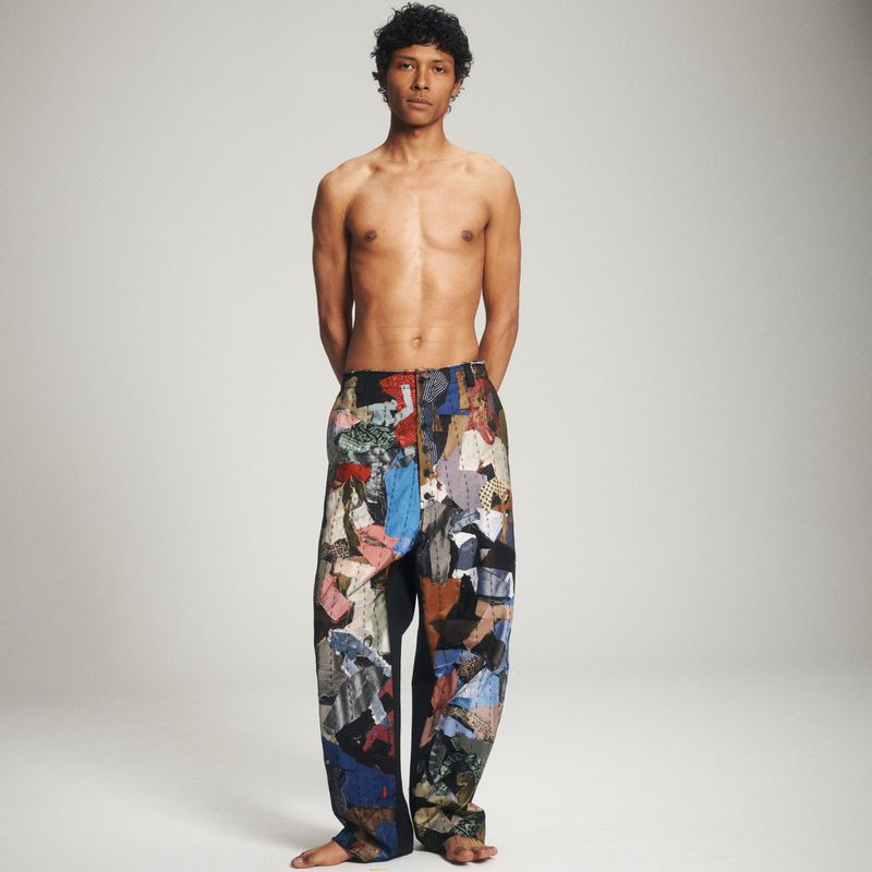Full-length portrait of a shirtless adult male wearing patchwork pants, standing in a neutral-toned studio, looking directly at the camera.