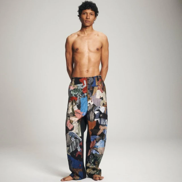 Full-length portrait of a shirtless adult male wearing patchwork pants, standing in a neutral-toned studio, looking directly at the camera.