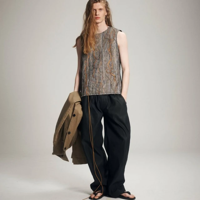 A slender man with long hair poses in an editorial fashion shot, wearing a sleeveless patterned top and dark pants, holding a beige jacket.