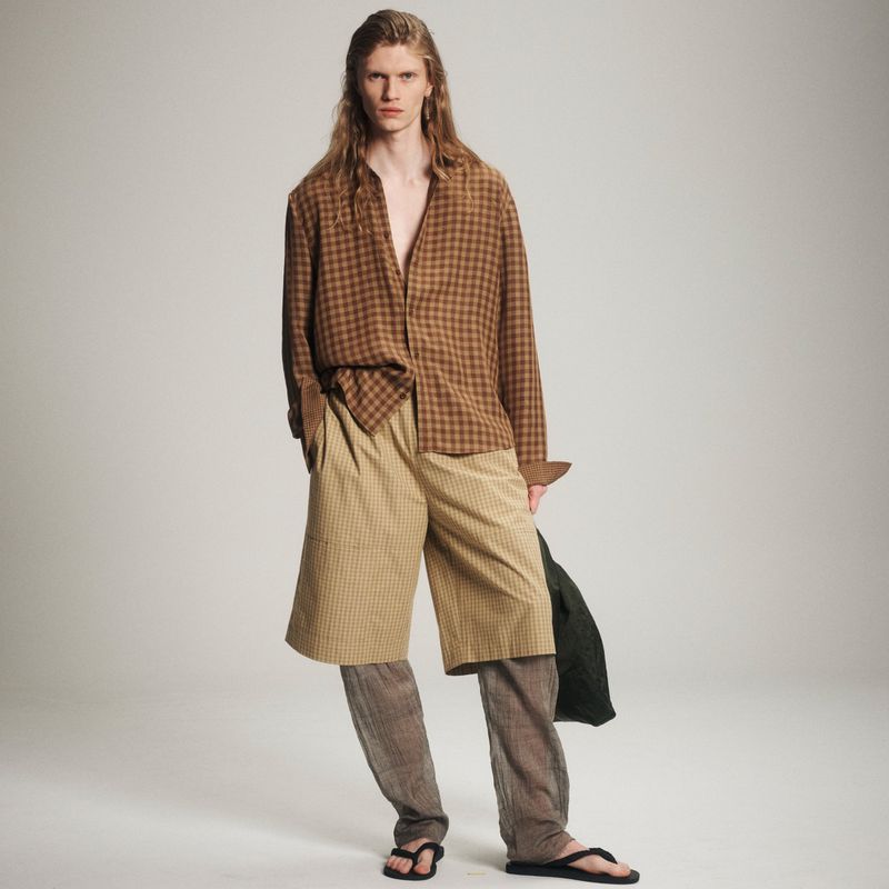 A person with long hair models a brown checked shirt, tan shorts, and black sandals against a neutral backdrop, showcasing a casual fashion style.
