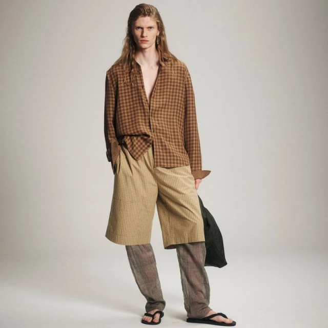 A person with long hair models a brown checked shirt, tan shorts, and black sandals against a neutral backdrop, showcasing a casual fashion style.