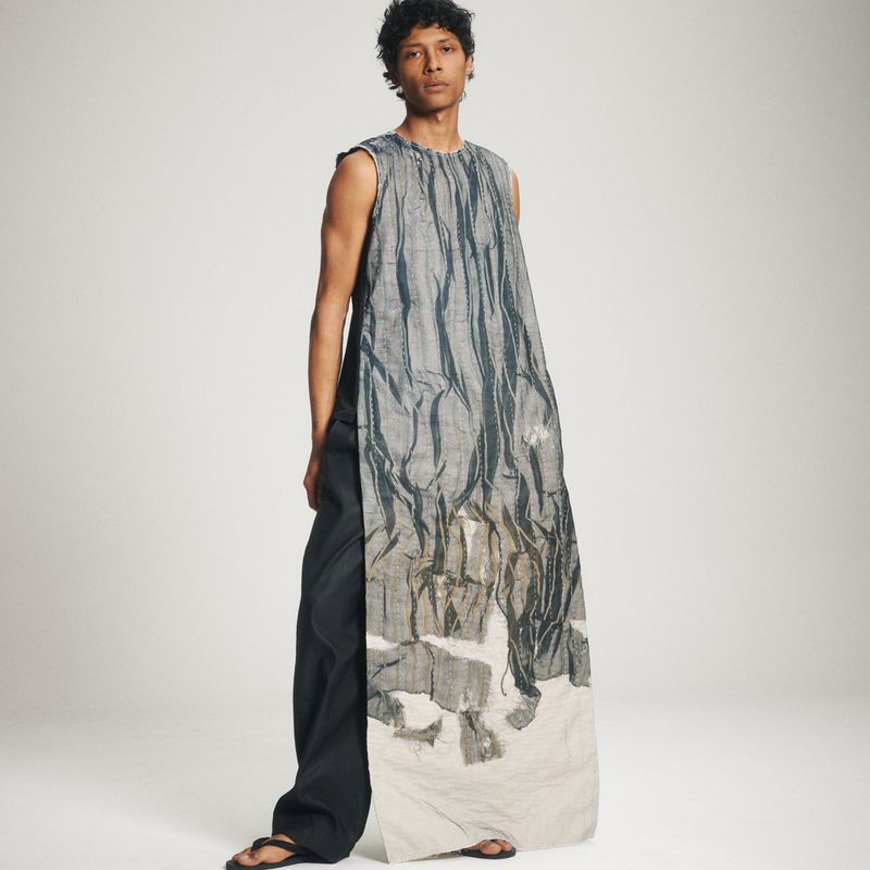 A male model wearing an elongated printed sleeveless tunic and dark wide-leg trousers against an off-white seamless studio backdrop.