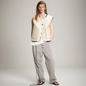 Model wearing a cream quilted vest and patterned grey trousers standing against a seamless grey backdrop.