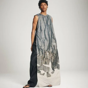 A male model wearing an elongated printed sleeveless tunic and dark wide-leg trousers against an off-white seamless studio backdrop.